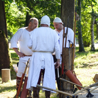 Three men dressed in medieval white tunics and caps stand in a wooded area at Dunamunde, engaged in conversation. Various medieval tools and weapons, including an axe and sword, are displayed on a wooden stand nearby. The scene is bright and sunny, with green trees providing a backdrop reminiscent of 2010.