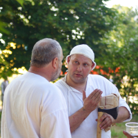 Two men in historical attire are conversing outdoors in Dunamunde. Both wear white tunics; the man on the right also wears a white head covering and holds an axe with a cloth on the blade. They stand in a green, leafy setting, seemingly engaged in discussion. A blurred person is in the background, hinting at 2010 reenactment activities.