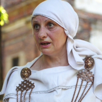 A woman wearing a white fabric headscarf and a white garment adorned with decorative bronze accents and chains stands in front of an out-of-focus background of a brick building. She appears to be speaking or reacting to something with a concerned or focused expression, reminiscent of the Dunamunde 2010 era.