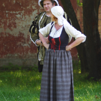 A man and woman dressed in historical costumes stand outdoors on grass, with a red and brown faded building in the background. The man, wearing an ornate outfit with a hat, and the woman, donning a long plaid skirt, white blouse, and headscarf, appear to be engaged in conversation or posing. Perhaps this scene is from the 2010 reenactment of Dunamunde.