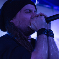 A vocalist with long hair wearing a black beanie passionately sings into a microphone on stage, embodying the spirit of folk metal reminiscent of Eluveitie. He has rings on his fingers and black wristbands on both wrists. The background features bright blue lights, creating a vibrant concert atmosphere. His eyes are closed, emphasizing his emotional performance.