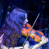 A woman with long, dark hair plays the violin under blue stage lighting. She wears a black sleeveless top, and tattoos are visible on her left arm. A drum set is partially visible in the background, suggesting a live music performance setting reminiscent of Swiss folk metal band Eluveitie.
