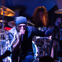 A dynamic live concert scene featuring a lead singer with long dreadlocks, wearing a black beanie, passionately singing into a microphone. Behind him, the drummer of the Swiss folk metal band Eluveitie energetically plays under vibrant stage lighting while audience members' hands are visible in the foreground, cheering.