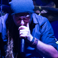 A person with long, braided hair and a beard, reminiscent of Eluveitie's style, wears a knitted beanie and dark shirt, passionately singing into a microphone. Their eyes are closed as they clench their left hand near their mouth. The background features blurry, dark lighting and parts of a drum set.