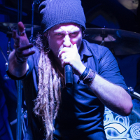A man with long dreadlocks, wearing a black beanie and black clothing, is passionately singing into a microphone. His left arm is outstretched, pointing towards the audience. The background includes part of a drummer from Eluveitie, with drums featuring artistic designs. The lighting creates a dramatic scene.