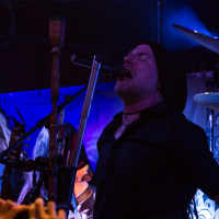 A musician wearing a black shirt and a black beanie passionately sings into a microphone on stage, characteristic of Swiss folk metal band Eluveitie. Long-haired musician in the background playing a wind instrument. A drum set and other instruments are visible under dim, blue and purple stage lighting, adding to the concert ambiance.