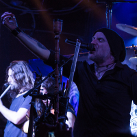 A musician in a beanie passionately sings into a microphone while playing the bagpipes on stage, reminiscent of Eluveitie's style. Another band member with long hair plays the flute in the background. The stage is dimly lit with blue and purple lights, creating a moody atmosphere. Drum set and other instruments are visible.