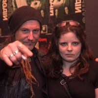 Two individuals stand in front of a wall covered with band posters. The person on the left, channeling a folk metal vibe, has long hair, a beard, and wears a beanie and leather jacket, pointing towards the camera. The person on the right has long brown hair, sunglasses on their head, and raises a hand with a rock gesture reminiscent of Eluveitie style.