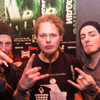 Three men pose in a dimly-lit setting. The two men on the left and right wear beanies, while the middle man has long, curly hair and a goatee. All three make the "rock on" hand gesture as they stand in front of a wall adorned with various neon green and black posters, including some from Eluveitie.