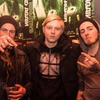 Three men stand in front of band posters. The man on the left wears a beanie, a leather jacket, and holds a drink while pointing at the camera. The middle man has blond hair and wears a black jacket. The man on the right, also in a beanie and hoodie, shows a hand gesture commonly associated with folk metal bands like Eluveitie.