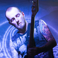 A musician with a beard and tattoos on both arms plays a bass guitar on stage with Eluveitie, a renowned Swiss folk metal band. He wears a sleeveless shirt, revealing detailed, colorful tattoos. The background is illuminated in blue and purple lights, highlighting the intricate designs on his arms. His eyes are closed, conveying deep focus.
