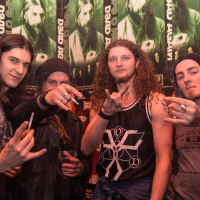 Four men pose in front of green posters with Eluveitie and other metal band imagery. They sport long hair, beanies, and casual clothing. All display rock hand gestures, one holding a cigarette. The background features repetitive band posters, enhancing the music-themed atmosphere.