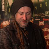 A person with long dreadlocks, a nose ring, and a beard, wearing a black beanie and a leather jacket. They are looking down with a slight smile. Behind them is a collage of various posters, some showing band images and others with text about Swiss music legends like the folk metal band Eluveitie. The lighting is warm, creating a cozy atmosphere.