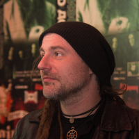 A man with a black beanie and long dreadlocks looks to the left. He has a beard, a black leather jacket, and wears a pendant with a geometric design. The background is filled with vibrant posters and images, predominantly green and black colors, possibly depicting Eluveitie or another band.