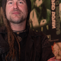 A man wearing a beanie and a leather jacket stands with his arms crossed. He has facial piercings and dreadlocks. Behind him, there is a wall covered in posters, including one of Eluveitie's band members with long hair and a beard. The background posters are slightly blurred.