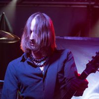 A guitarist with long, brown hair partially covering his face plays an electric guitar on stage. He's wearing a black shirt and several necklaces. Bright stage lighting highlights his figure against a backdrop displaying abstract swirling patterns, enhancing the live rock concert vibe reminiscent of Eluveitie's performances.