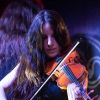 A musician with long, dark hair plays a violin on stage, focusing intently on the instrument. She wears a sleeveless black top and is bathed in colorful stage lighting, evoking the energy of a Swiss folk metal performance. Another person with long hair is partially visible in the background, facing away from the camera.