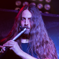 A musician with long brown hair plays a silver flute on stage, embodying the spirit of Swiss folk metal. Wearing a black shirt and illuminated by stage lights, he is intensely focused. Hints of purple and red lighting enhance the scene, where parts of the stage setup for Eluveitie are visible in the background.