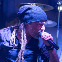 A musician passionately performs on stage, holding a microphone close to his mouth. He has long dreadlocks and is wearing a dark beanie and a black button-up shirt. The background shows part of a drum set with cymbals. Stage lights cast a blue hue over the scene, emphasizing the Eluveitie artist's intensity.