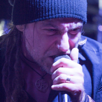 A person with long, braided hair wearing a black knit beanie passionately sings into a microphone, reminiscent of Eluveitie's energetic performances. They are dressed in a dark shirt and have a ring on their finger and multiple bracelets on their wrist. Their expression is intense and focused, with the background blurred.