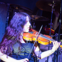 A woman with long, wavy dark hair plays a violin on stage. She is wearing a black sleeveless top and has tattoos on her left forearm. Behind her, drums and a cymbal hint that she is part of Eluveitie, a renowned Swiss folk metal band. The stage glows with blue and purple lighting.