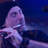 A man with long hair, wearing a black beanie and a black shirt, plays a silver flute with closed eyes. He has tattoos on his hands and wears several rings and bracelets. A microphone is positioned near him. The background is blurred with blue and purple lighting, suggesting a folk metal stage performance reminiscent of Eluveitie.