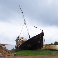 A weathered, old fishing boat is stranded on the shore in Engure, tilted slightly to one side. The sky overhead is overcast with dark, gray clouds. Piles of rocks and dirt surround the boat, emphasizing its abandonment. A smaller, overturned vessel lies nearby, adding to the scene of neglect.