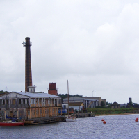 An old industrial building with a tall chimney stands by the calm Engure river, under an overcast sky; a vintage boat moored nearby and buoy markers floating on the water.