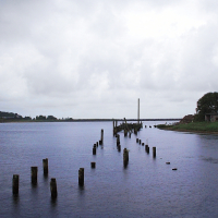 A serene, overcast river scene in Engure with remnants of an old pier jutting out into the water, a rural landscape and buildings on the riverbank under a moody sky.