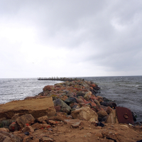 A rugged, rocky pier in Engure juts into a tumultuous sea under a gloomy sky, with colorful boulders forming a barrier against the waves.