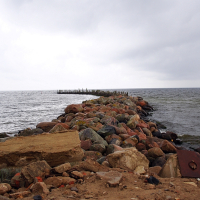 A rocky breakwater extends into the sea at Engure, comprised of large, multicolored stones. The water is calm, with gentle waves lapping against the rocks. The sky is overcast with thick, gray clouds. In the foreground, additional stones and some debris rest on sandy ground.