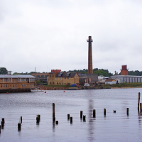 A tranquil overcast day by the riverside in Engure, featuring an industrial landscape with a prominent chimney, older buildings, and mooring posts in the foreground, reflective of a bygone era's