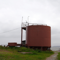 A large, rusty cylindrical tank with a metal staircase stands under an overcast sky near the Engure body of water, conveying a sense of industrial abandonment and solitude.