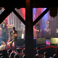 At the 2011 Fontaine Festival, a rock band performs on stage with vibrant red and blue lighting. The lead singer, wearing a black hat, engages with the audience while the guitarist and bassist play alongside. A wooden beam frames the stage. An audience watches the performance, and a photographer captures the moment.