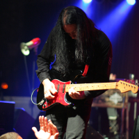 A guitarist with long black hair, dressed in black, is passionately playing an electric guitar on stage during the 2011 Fontaine Festival. He is leaning forward towards the audience, who are reaching out towards him. The stage is dimly lit with blue and purple lights, creating a vibrant atmosphere.