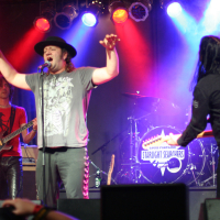 At the 2011 Fontaine Festival, a man in a hat and graphic t-shirt sings passionately into a microphone on stage, with arms raised. Another musician to the left plays an electric guitar, and a third man to the right readies another instrument. The backdrop features colorful lights and a drum set with "Starlight Searchers" written on it.