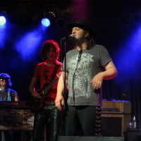 At the 2011 Fontaine Festival, a rock band performs on stage under blue and purple lighting. The lead singer, wearing a black hat and gray t-shirt, sings into a microphone. To his left, a guitarist with curly hair and a red shirt plays. A keyboardist and drum set are visible in the background.