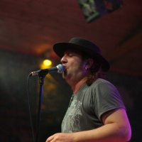 A person with long hair wearing a black hat and a gray T-shirt sings into a microphone on a dark stage at the Fontaine 2011 festival. They are bathed in a mix of stage lights, including a focused yellow light above their head. The background is dimly lit, with a visible light rig hanging from the wooden ceiling.