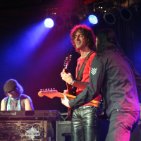 Three musicians perform on stage at the Fontaine festival under colorful lighting. One plays a keyboard on the left, wearing a hat. The second musician, in the middle, plays an electric guitar and is smiling. The third musician, on the right, faces away from the camera, playing another electric guitar.
