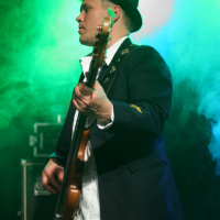 A musician in a black fedora and dark blazer plays a guitar on stage at the 2011 Fontaine festival. He stands under green and blue lights, partially obscured by stage smoke. The musician looks focused, with his gaze directed to the side. The background shows stage equipment in soft focus.