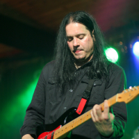 At the 2011 Fontaine festival, a musician with long black hair plays an electric guitar on stage. He is wearing a black shirt and is focused on his performance. The guitar has a red strap, and green and blue stage lights in the background create a vibrant atmosphere.