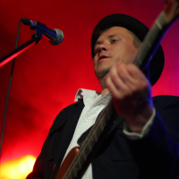 A musician wearing a black hat and a black jacket over a white shirt plays an electric guitar on stage at the 2011 festival. The background is illuminated with vibrant red and yellow lights, and a microphone is visible in front of the performer. The atmosphere suggests an energetic live music performance.