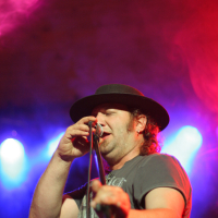 At the Fontaine Festival 2011, a person with a beard wearing a black hat and a gray t-shirt is singing into a microphone on stage, illuminated by vibrant red and blue stage lights. Their eyes are closed as they hold the microphone stand with their right hand, conveying emotion in their performance.