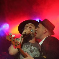 Two male musicians perform on stage amid vibrant, colorful lights at the Fontaine Festival 2011. One, wearing a black hat and graphic t-shirt, sings into a microphone. The other, in a suit and dark hat, leans in close, holding a guitar and also singing. The background is illuminated with orange, red, and purple lighting.