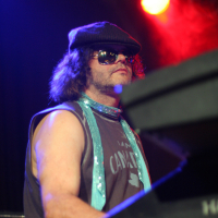 At the 2011 Fontaine Festival, a person plays a keyboard on stage, illuminated by red and blue lights. They wear a black cap, sunglasses, and a sleeveless dark shirt. A light blue, glittery scarf is draped around their neck. The background is dark, highlighting the individual and creating a dramatic atmosphere.