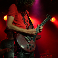 A guitarist with long curly hair, wearing a red shirt and black pants, plays an electric guitar under red and purple stage lights at the Fontaine Festival 2011. Another person stands in the background, partially visible. The stage is dimly lit with a blurred audience in the background.