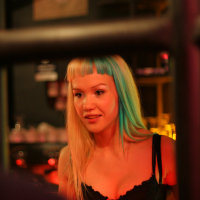 A woman with two-toned straight hair, blonde and green, is photographed indoors under warm, dim lighting. She has blunt bangs and is wearing a black top with a pink bow. In the slightly blurred background featuring decor elements and various bar items, a poster for the Fontaine festival 2011 can be seen.