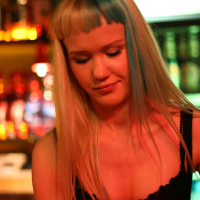 A person with long blonde hair featuring blue highlights and blunt-cut bangs is looking down and slightly smiling. They are wearing a black top with a red ribbon. The background is illuminated with warm, colorful lights, suggesting a lively indoor setting like the 2011 Fontaine Festival or a bar.