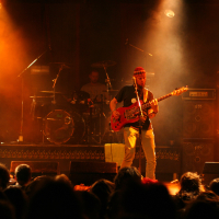 A guitarist performs on stage under warm amber lighting during the Fontaine Festival 2011, wearing a hat and casual clothing. A drummer is visible in the background, playing amidst smoke effects. Amplifiers and equipment are positioned around the stage. An audience watches from the foreground, silhouetted by the stage lights.