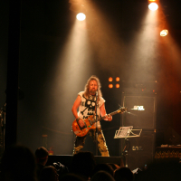 A guitarist with long hair is performing on stage under warm stage lights at the 2011 Fontaine Festival. He is wearing a white sleeveless shirt and camouflage pants, playing an electric guitar. Behind him, amplifiers and a music stand are visible. The audience is partially visible in the foreground, watching the performance.